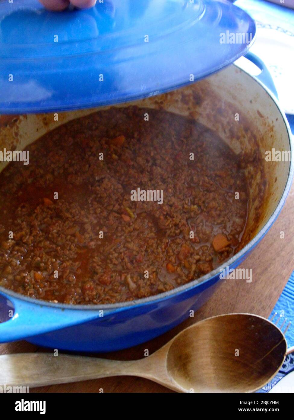 Raising the lid on a classic Italian bolognese meat sauce in large blue enamel casserole with big wooden spoon - Stock Photo