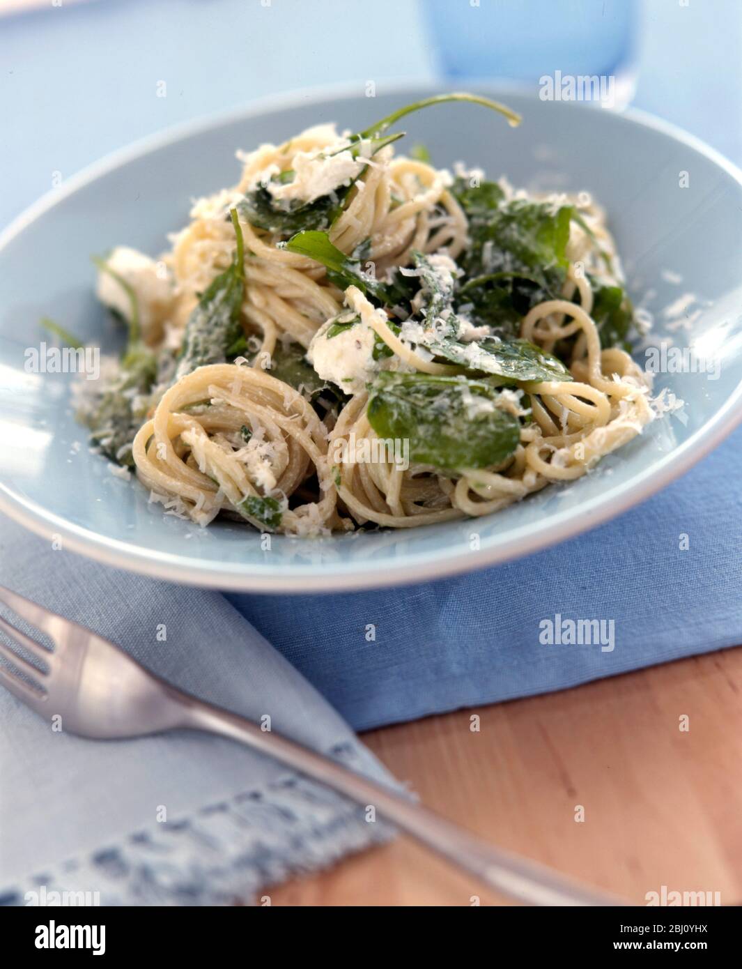 A pale blue ceramic bowl of spaghetti garnished with basil and grated parmesan on blue cloth - Stock Photo