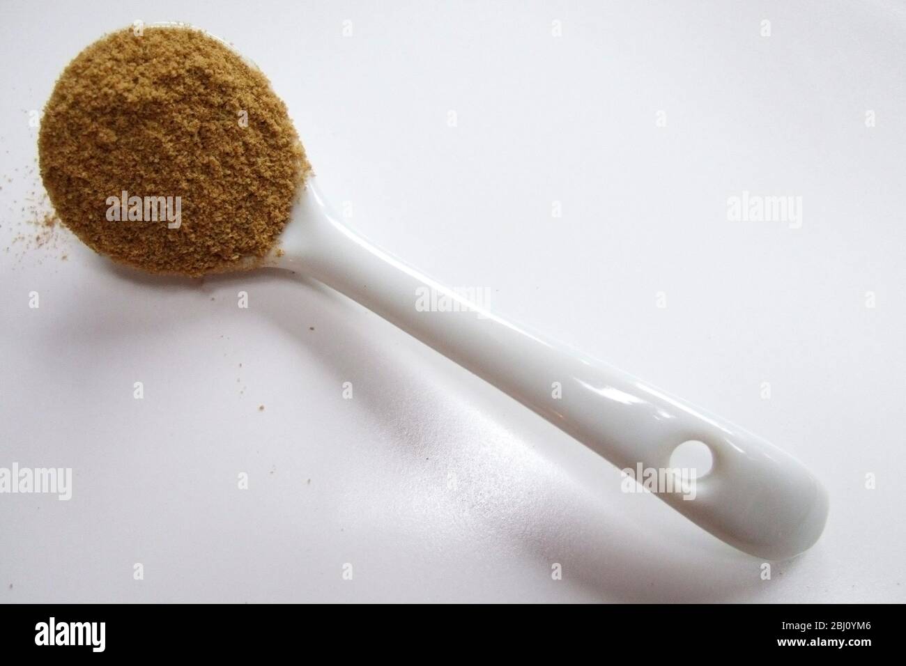 Porcelain spoon heaped with ground cumin powder on white ceramic surface - Stock Photo