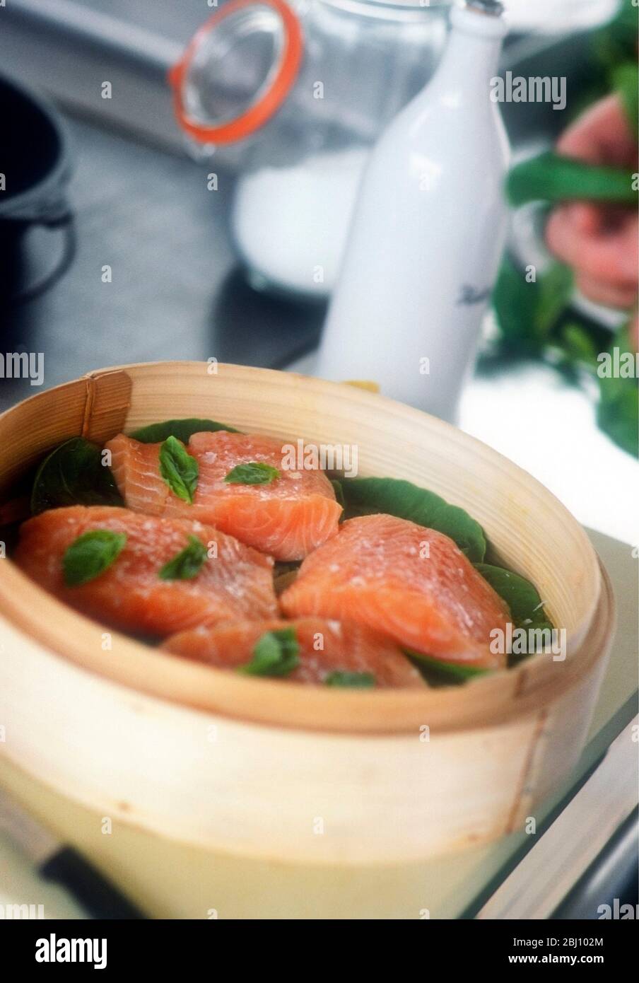 Salmon fillets set to steam on bed of spinach with basil leaves in kitchen setting - Stock Photo