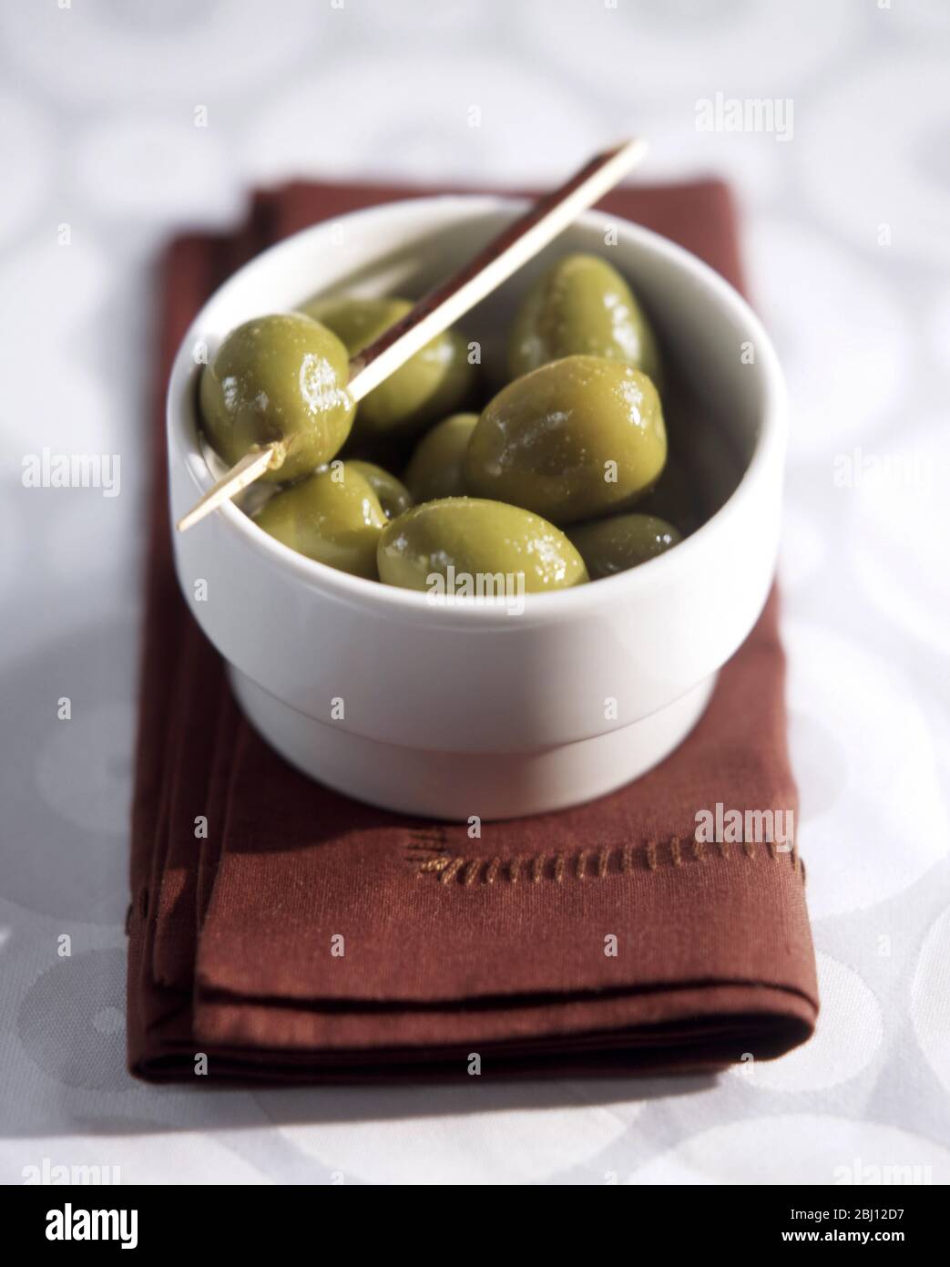 Small white dish of large queen olives on brown napkin with toothpick spearing one olive - Stock Photo