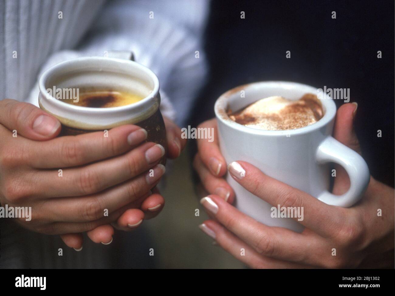 Hot drinks in hands - topped with frothy milk and chocolate powder - Stock Photo