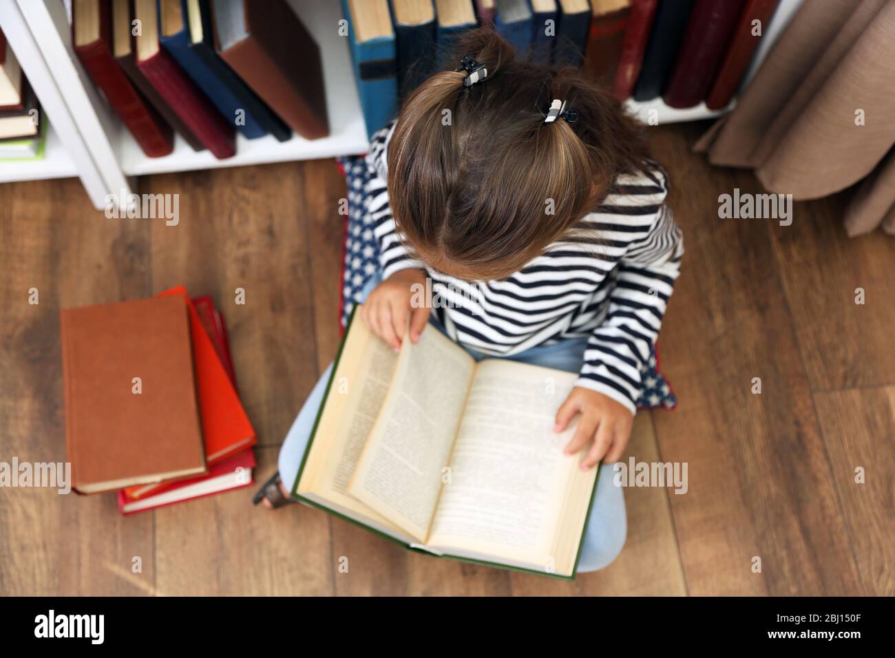 Beautiful little girl with books sitting on floor in library Stock ...