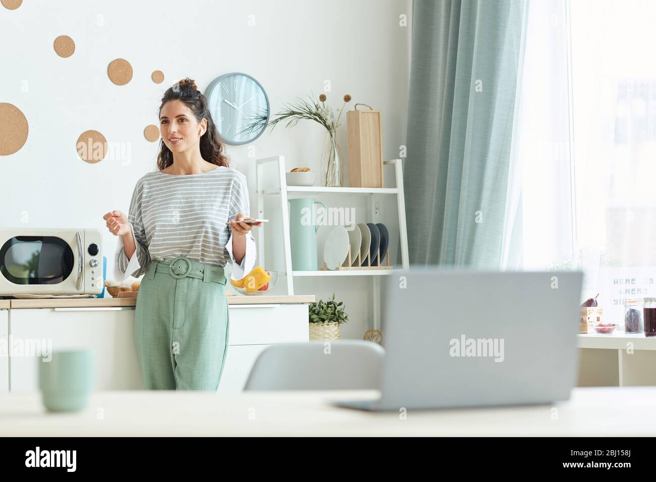 Young business person standing in her kitchen discussing something with her colleagues using online meeting application on smartphone Stock Photo