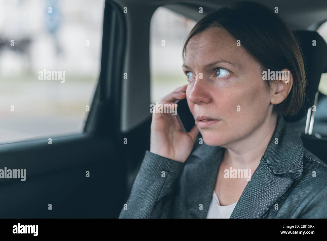 Surprised businesswoman talking on mobile phone on car back seat, business on the move concept. Elegant adult caucasian female person using smartphone Stock Photo