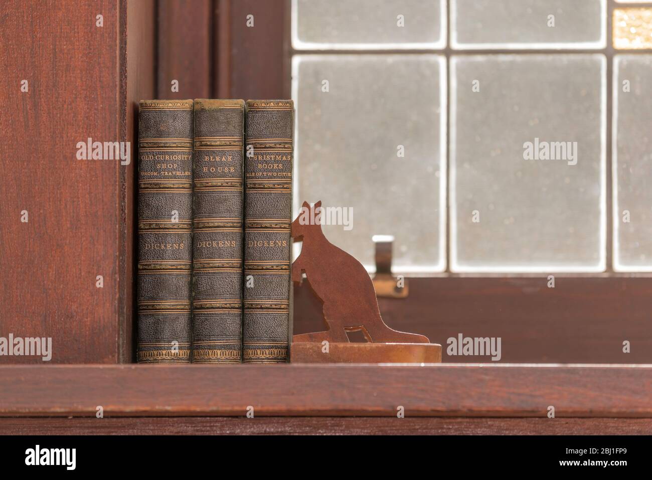 Three antique leather bound Charles Dickens books sitting in a timber window-sill held up by Australian kangaroo bookends with lead light background Stock Photo