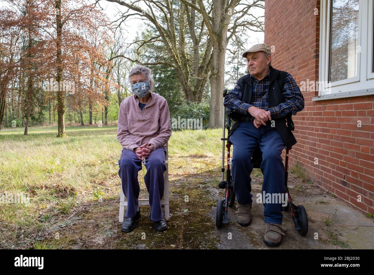 Seniorenpaar sitzt am Haus im Garten , aud einem Stuhl und einem Rollator. Die Seniorin trägt einen Mundschutz. Stock Photo