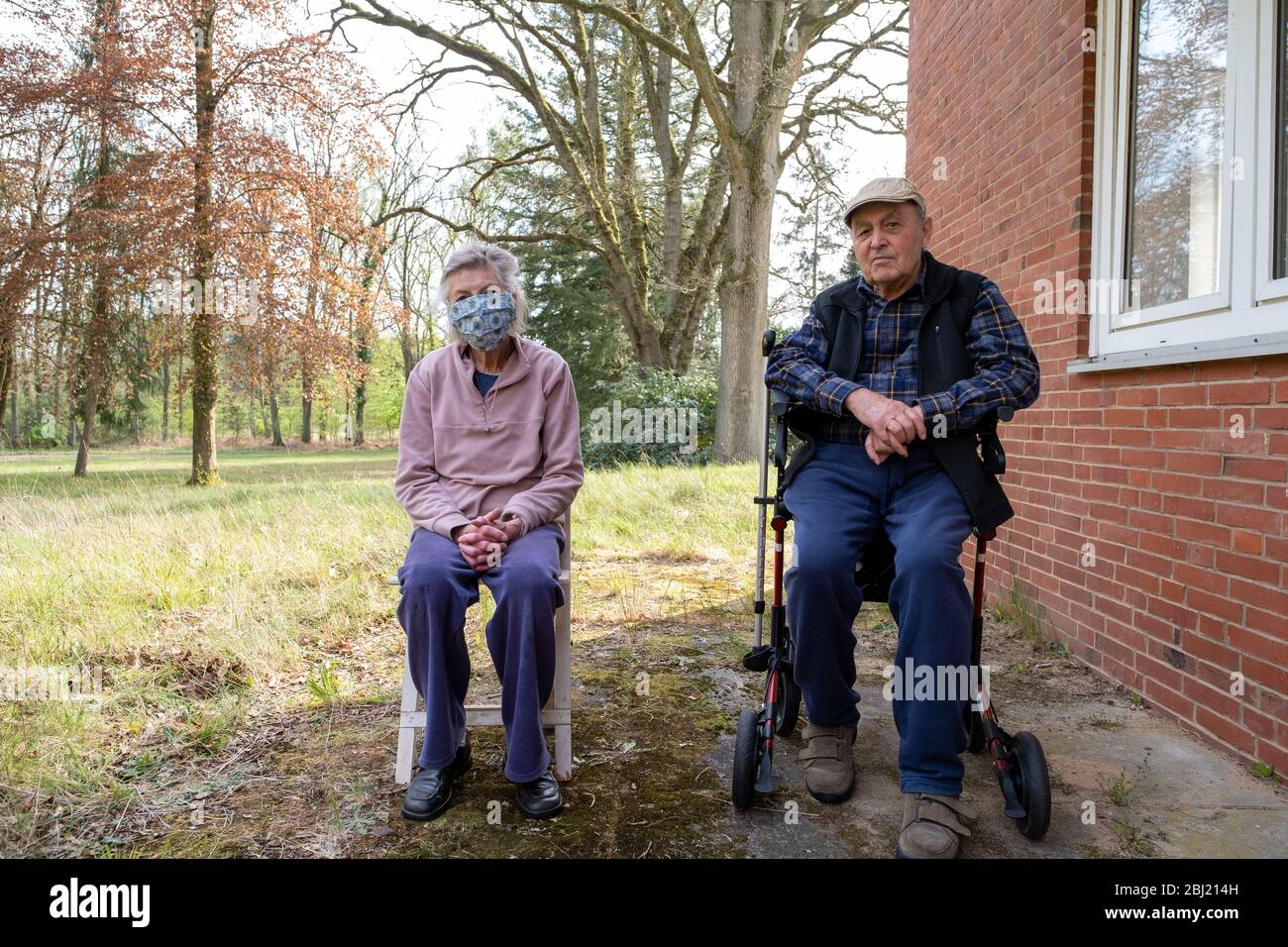 Seniorenpaar sitzt am Haus im Garten , aud einem Stuhl und einem Rollator. Die Seniorin trägt einen Mundschutz. Stock Photo