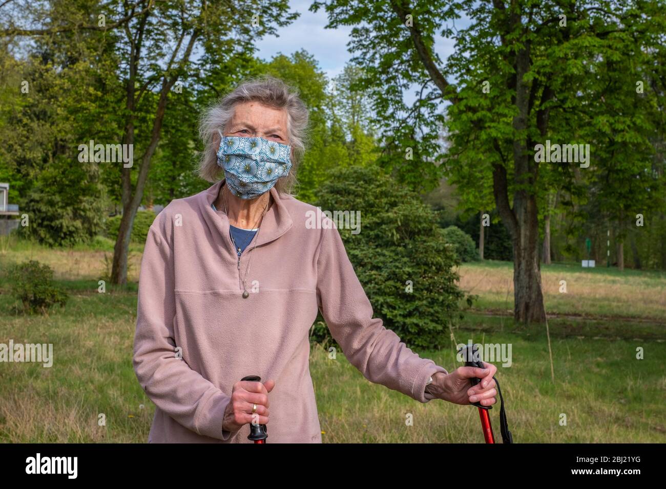 Seniorin mit Walking-Stöcken als Gehhilfe in der Natur mit Maske. Die Seniorin trägt einen Mundschutz. Stock Photo