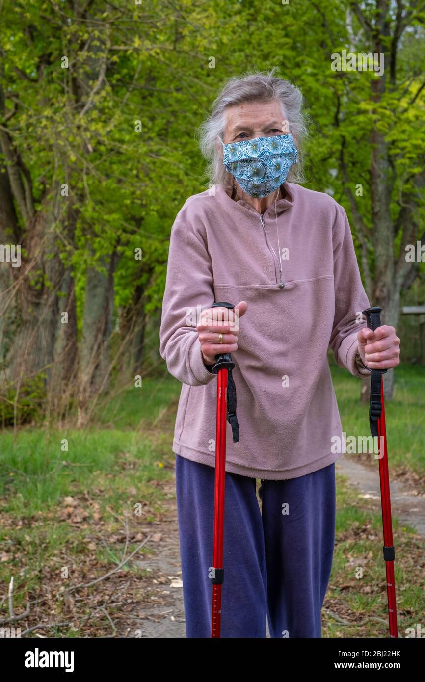 Seniorin mit Walking-Stöcken als Gehhilfe in der Natur mit Maske. Die Seniorin trägt einen Mundschutz. Stock Photo