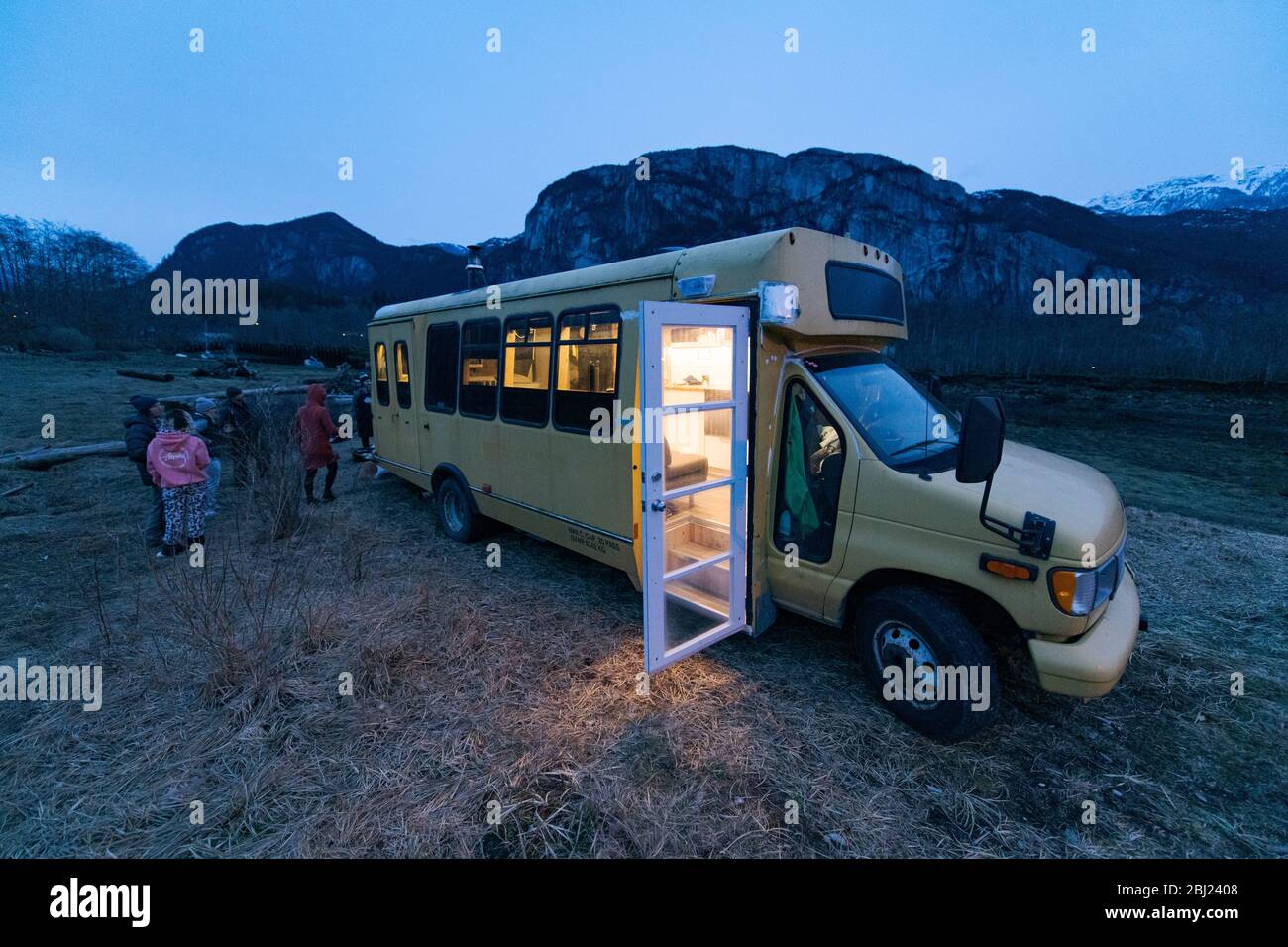 Campervan in a rocky landscape at dusk with the light on inside and the door open, group of people standing towards the rear. Stock Photo