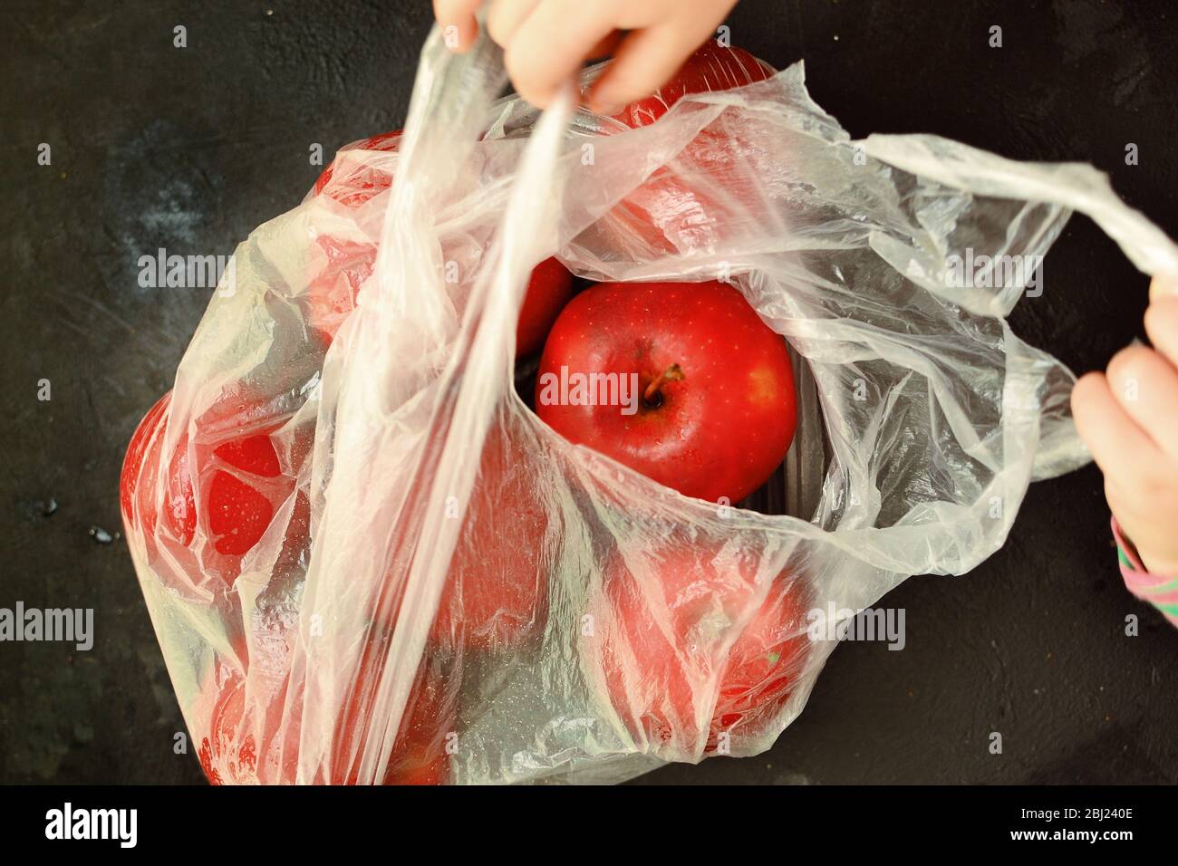 Red ripe apples in a plastic bag. Dark background, top view. Buying fruit. Hands of a child in the frame. The child holds and opens a bag of apples Stock Photo