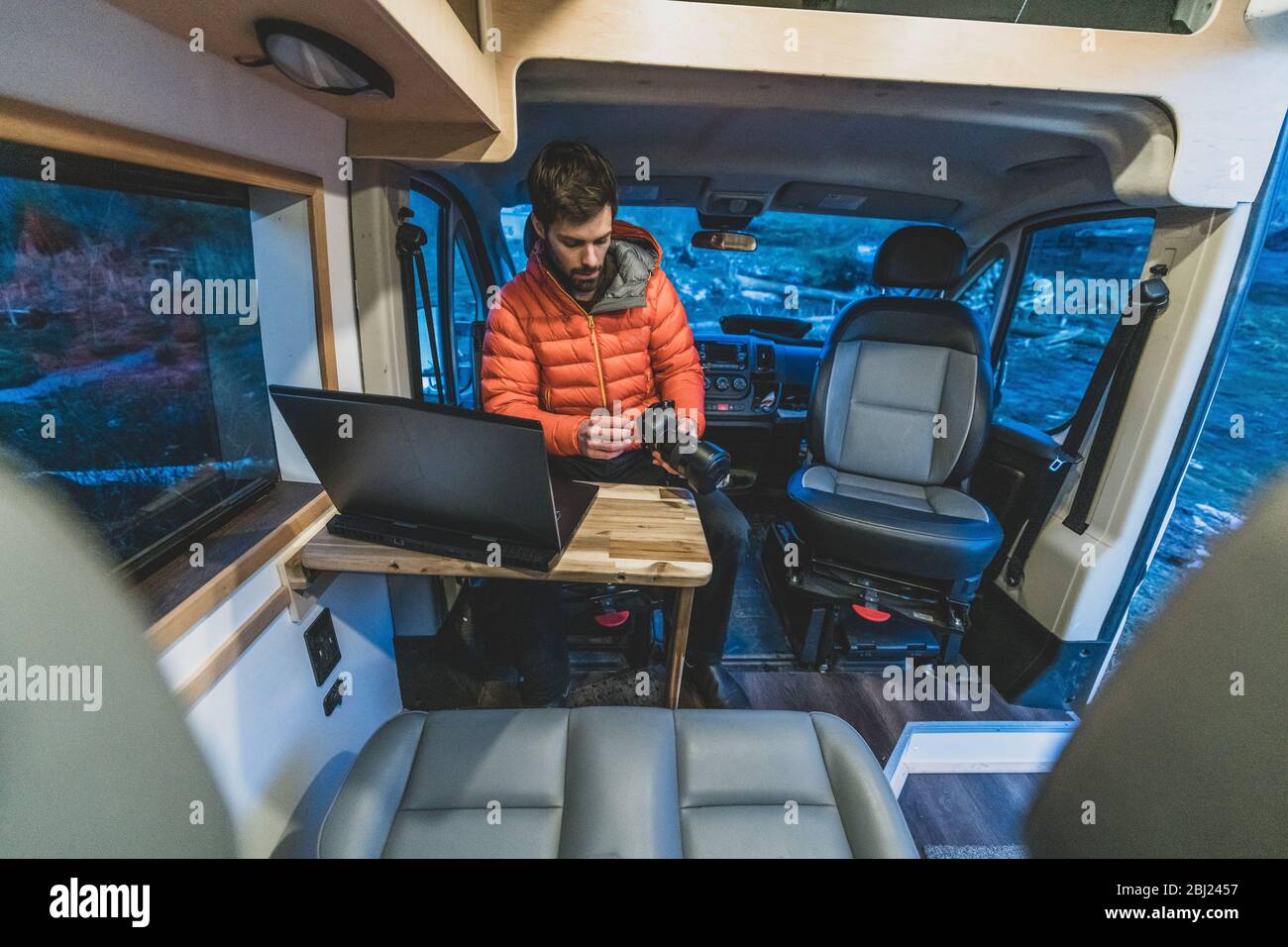 A man sitting in his campervan at a table with a laptop open looking at a camera. Stock Photo