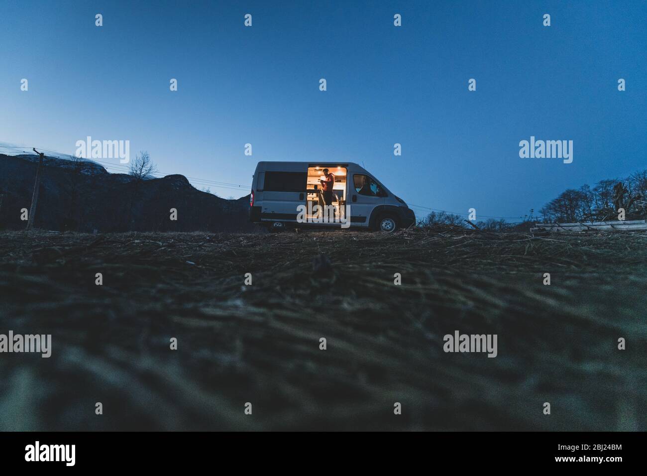 Campervan silhouetted on a hill in the evening with the lights on inside and a man standing by the open side door. Stock Photo