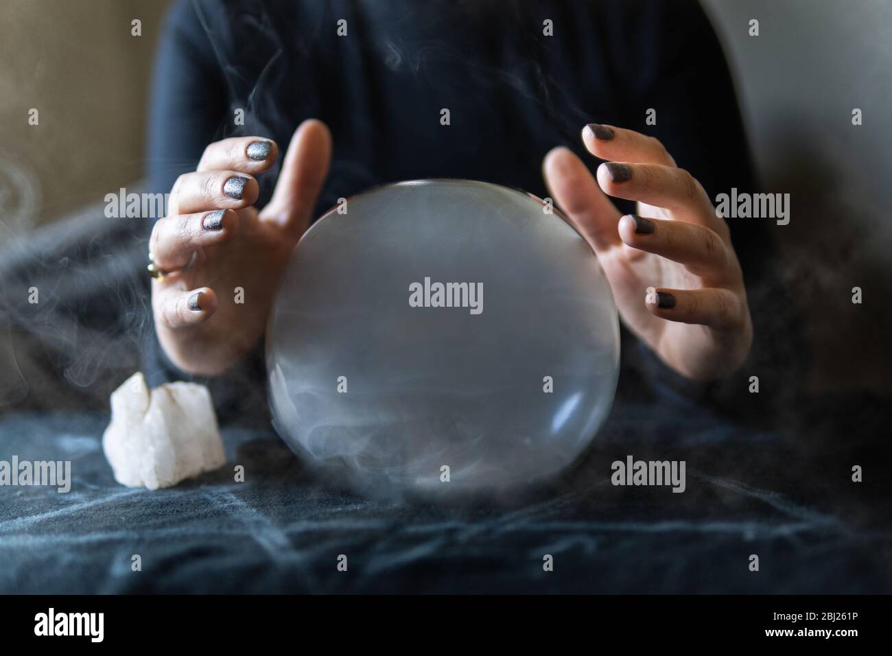 Fortune teller holding hands above magic crystal ball with smoke around. Conceptual image of practicing black magic and occultism. Stock Photo