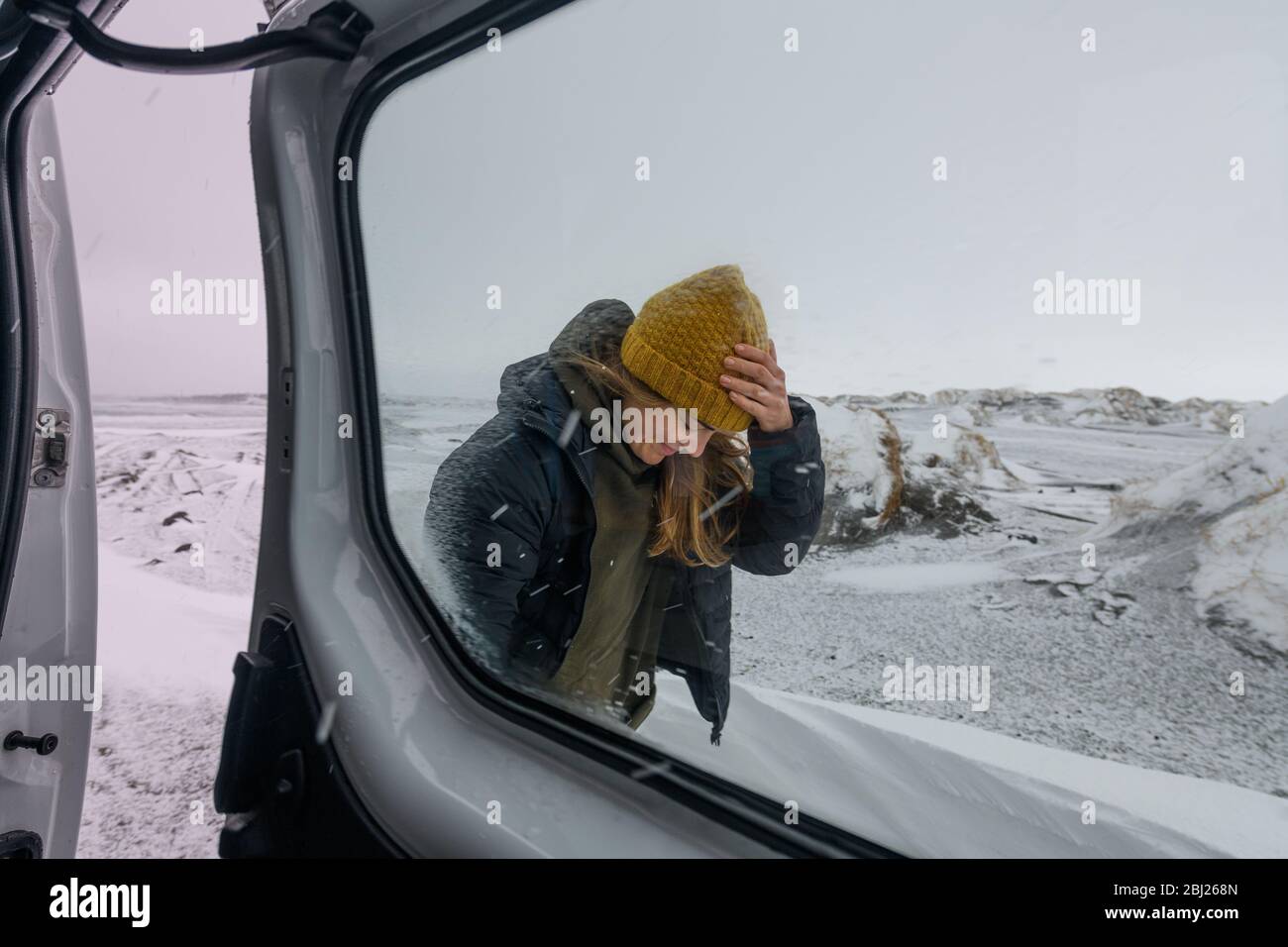 A woman wearing a woolly hat and jacket standing in a snowy landscape through a campervan window. Winter Surfing. Stock Photo