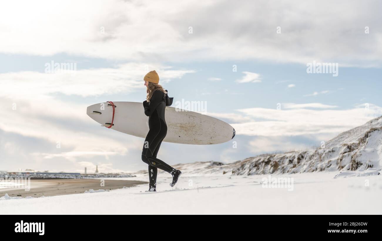 A woman wearing a wetsuit and holding a surfboard walking down a snowy beach and looking out to sea. Stock Photo
