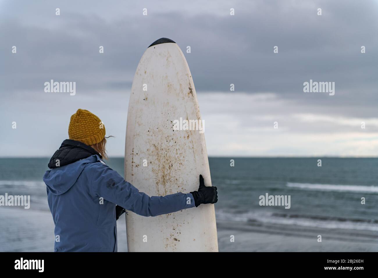 Rear view of a woman holding a surfboard standing on a snowy beach and looking out to sea. Stock Photo