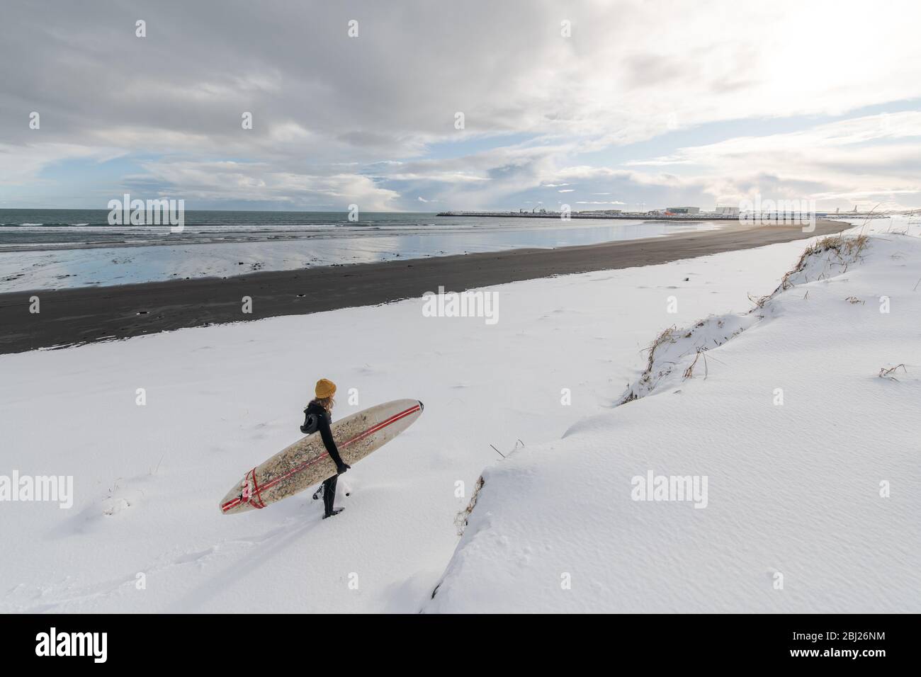 Rear view of a woman wearing a wetsuit and holding a surfboard walking along a snowy beach. Stock Photo