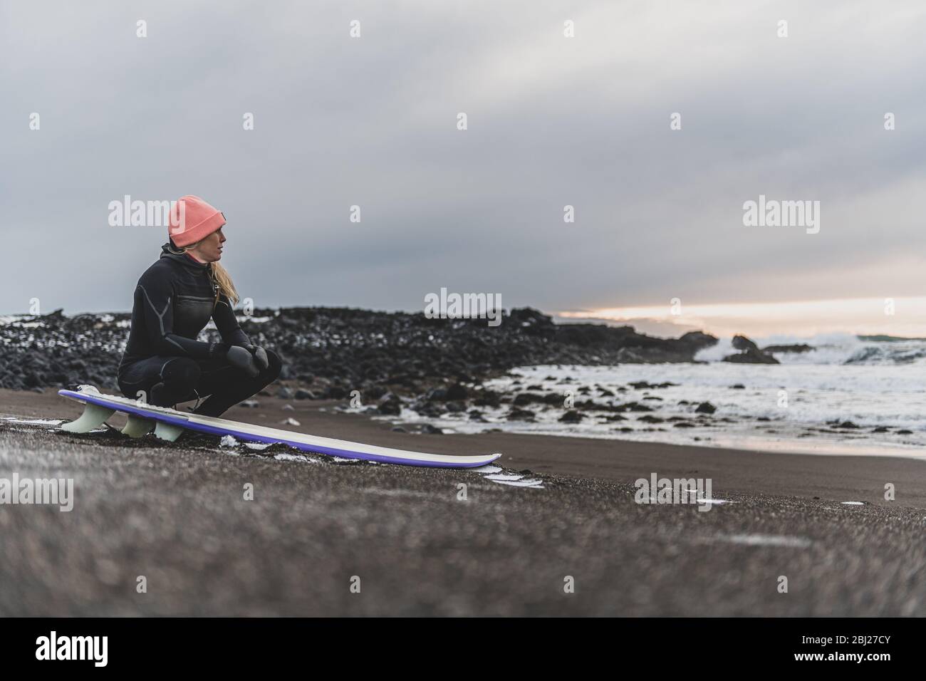 A woman sitting on her surfboard on a beach looking out to sea. Stock Photo