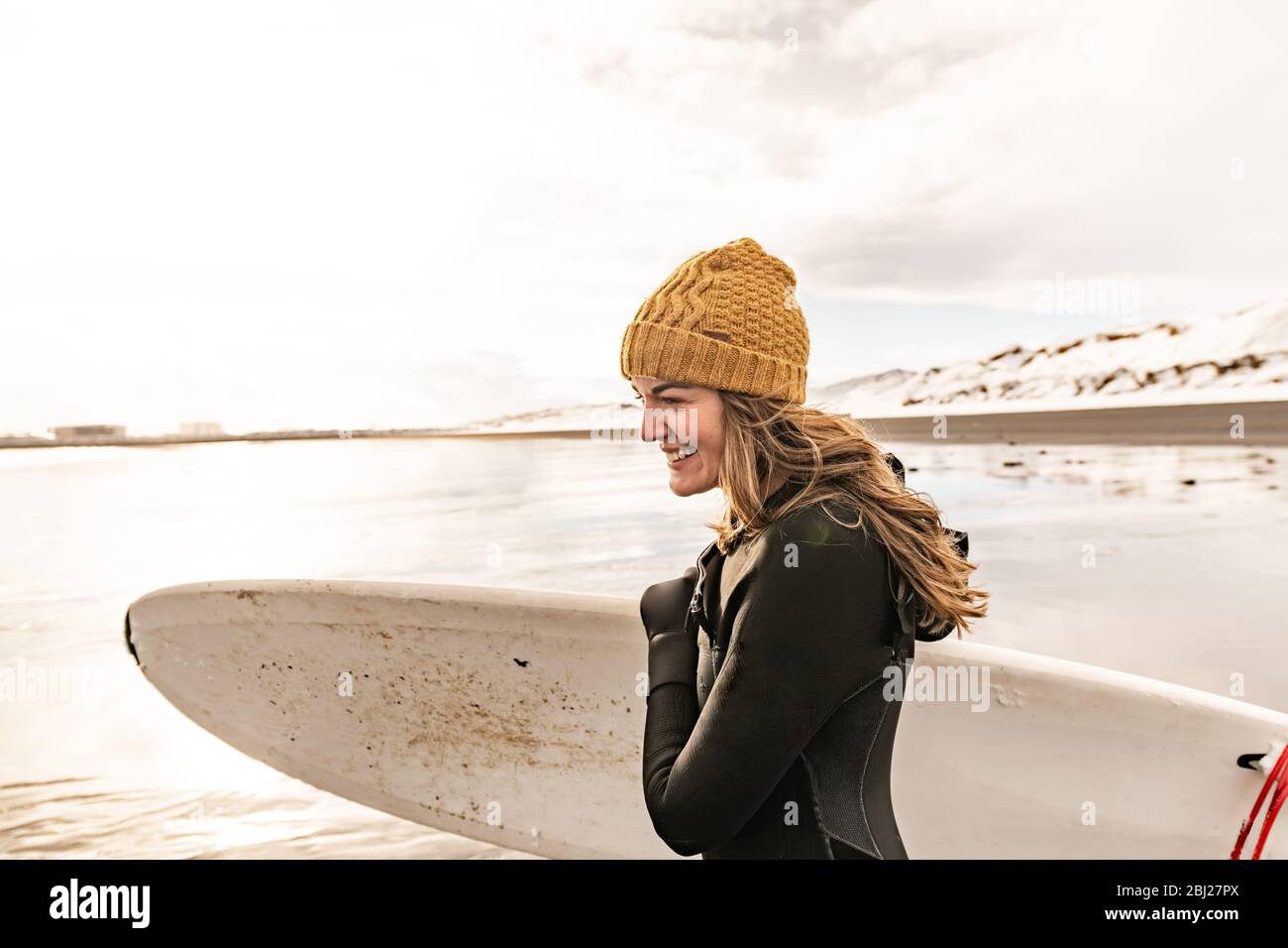 A woman wearing a wetsuit and woolly hat holding a surfboard on a beach with a snowy hill in the background. Stock Photo
