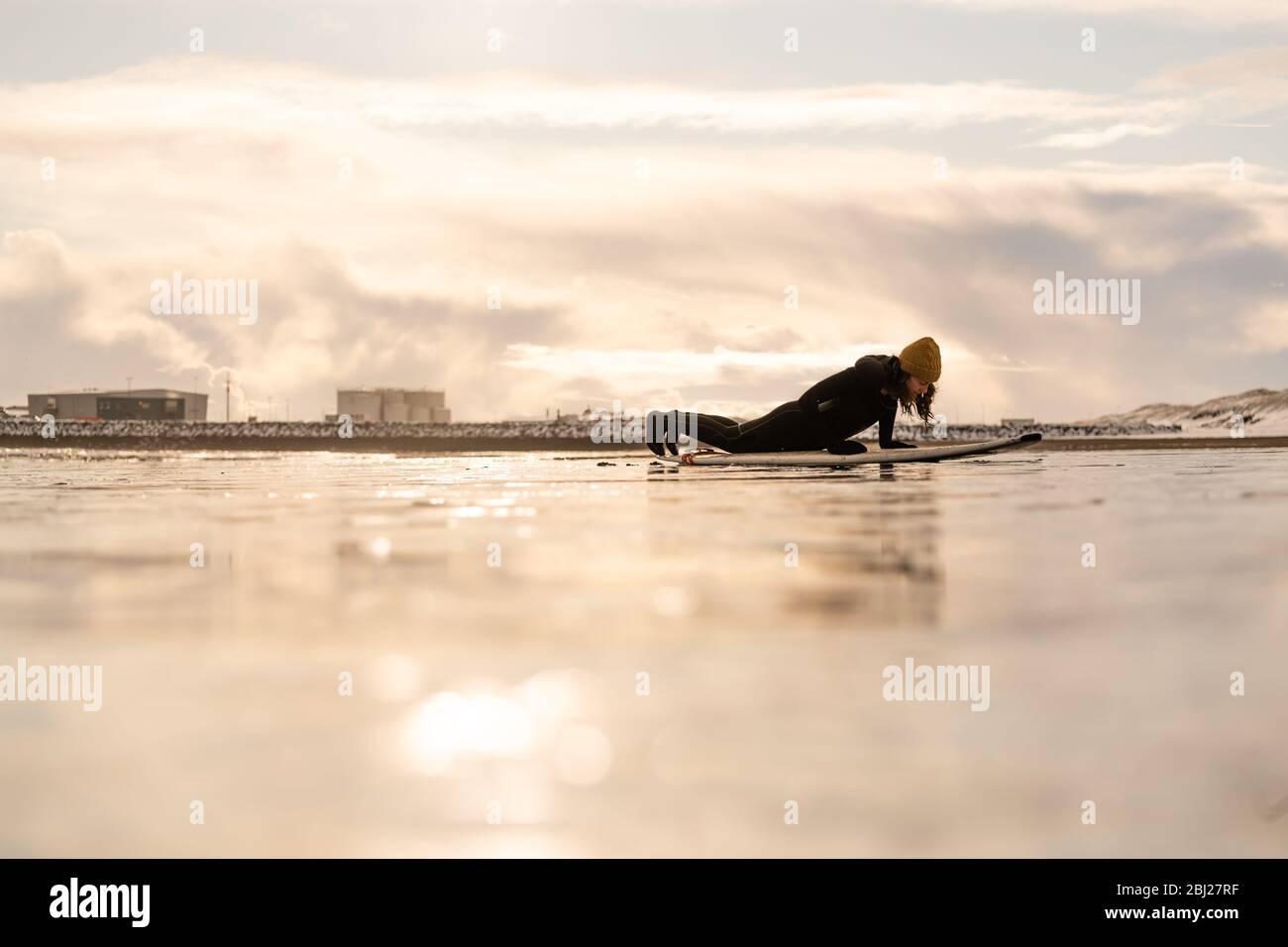 Silhouette of a woman with a surfboard lying on a beach with industrial units on the far horizon. Stock Photo