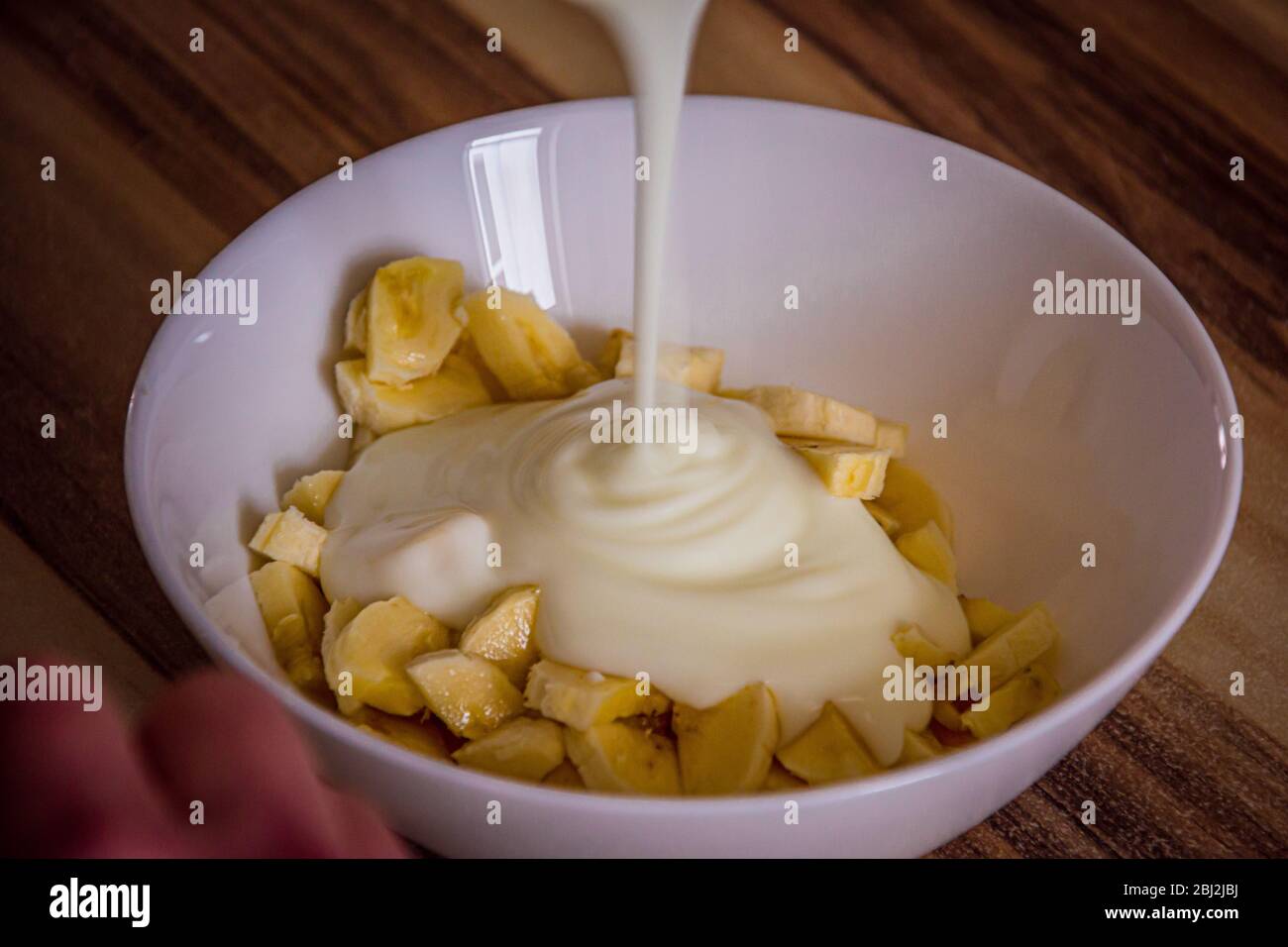 pouring natural yogurt on banana pieces in a white round bowl Stock Photo