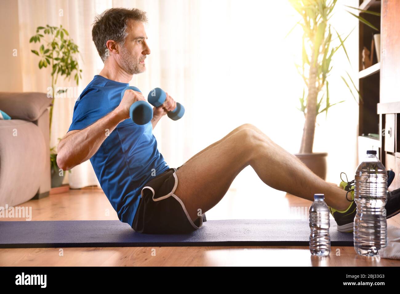 Man doing dumbbell exercises sitting on mat in his living room watching