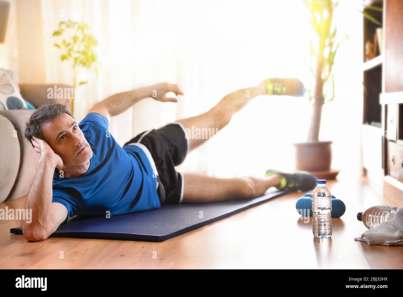 Man doing leg exercises lying on mat in his living room with water and dumbbells on the floor Stock Photo