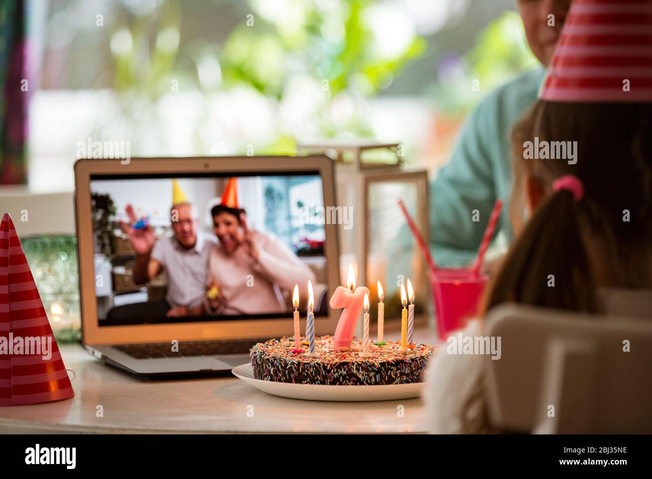 Happy little girl celebrating birthday at home with parents and grand parents on video call. Laptop with senior couple online, cake with candles Stock Photo