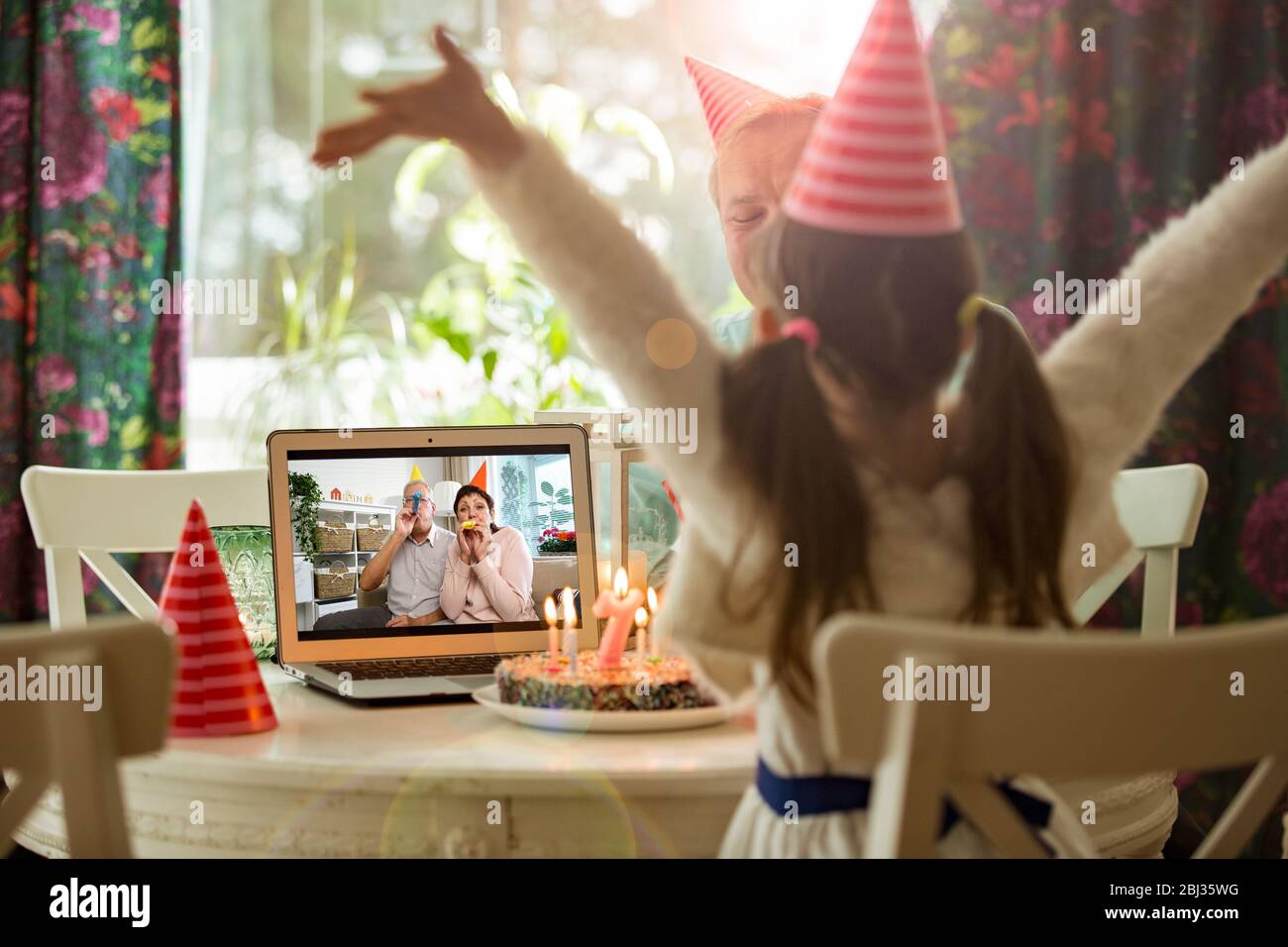 Happy little girl celebrating birthday at home with parents and grand parents on video call. Laptop with senior couple online, cake with candles Stock Photo