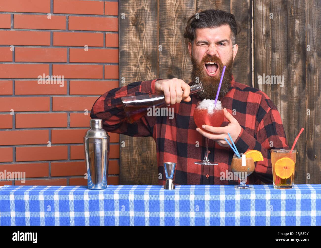 Barman with beard and shouting face makes cocktail, pouring glass with alcohol. Barman or hipster mixing drink. Man holds wineglass and bottle on wooden texture background. Making drinks concept. Stock Photo