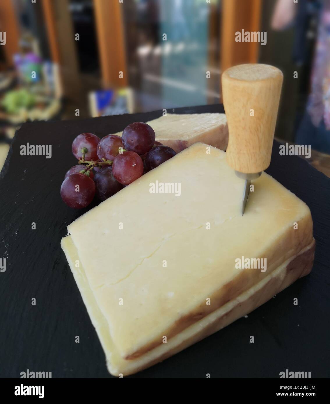 Big block of hard cheese stabbed with cheese cutting instrument or knife, on a wooden platter paired with red grapes ready for tasting and blurry back Stock Photo