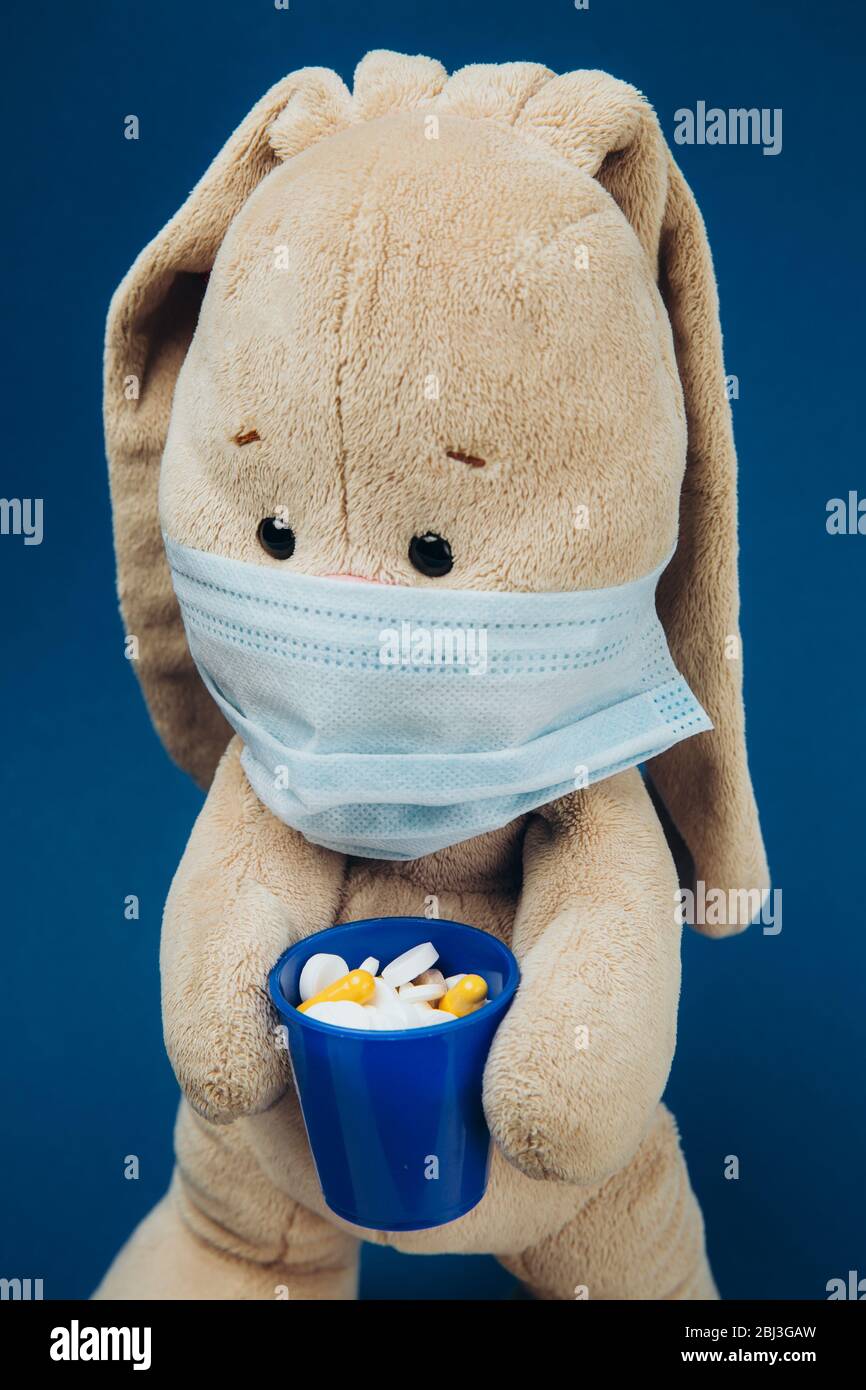The toy bunny wears a protective medical mask. Flapper holds a glass of pills. Stop coronavirus. Medicine concept Stock Photo