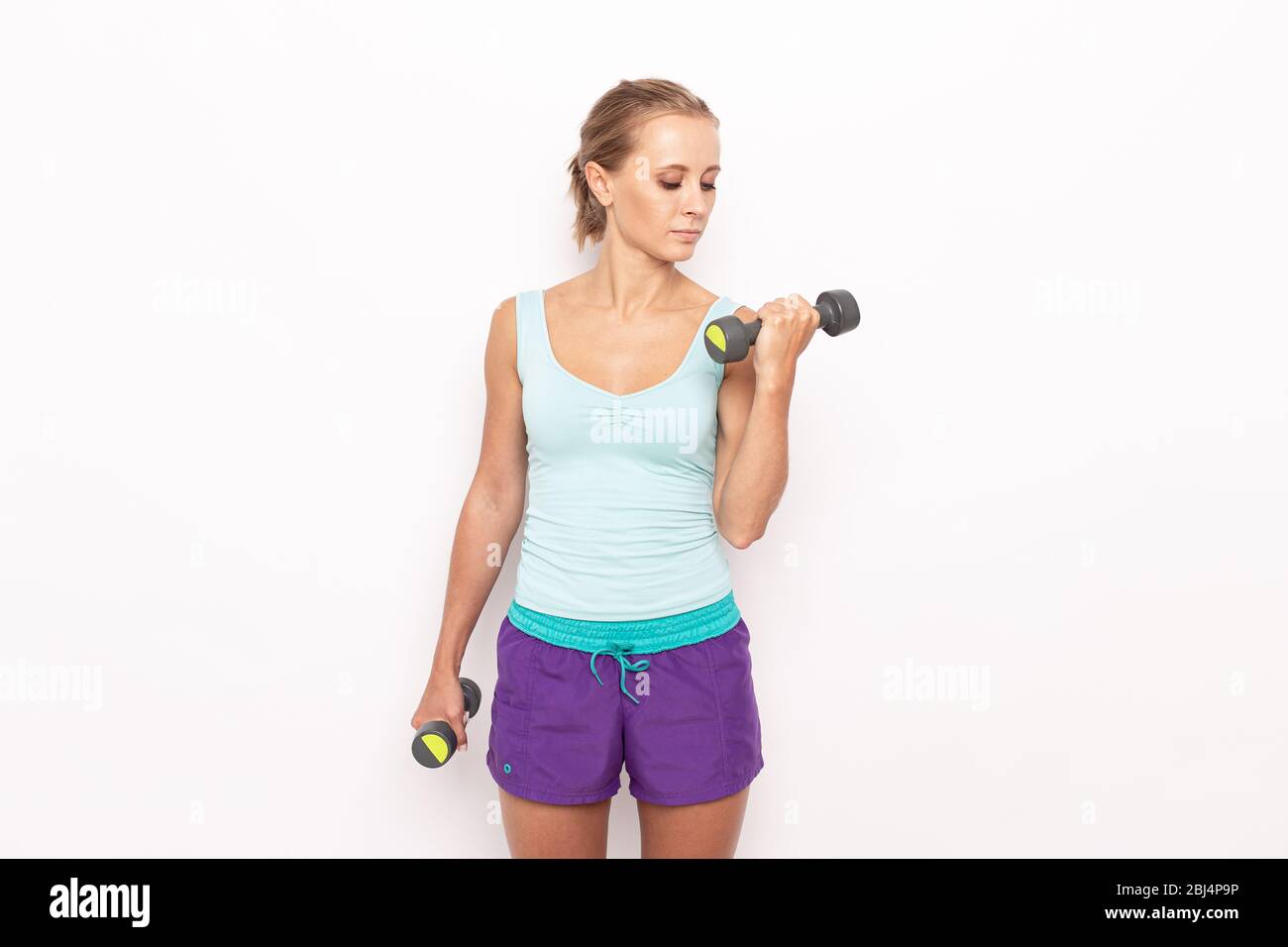 Confident-looking sportswoman raising left hand with dumbell while her right hands stay still. White isolated background. Stock Photo