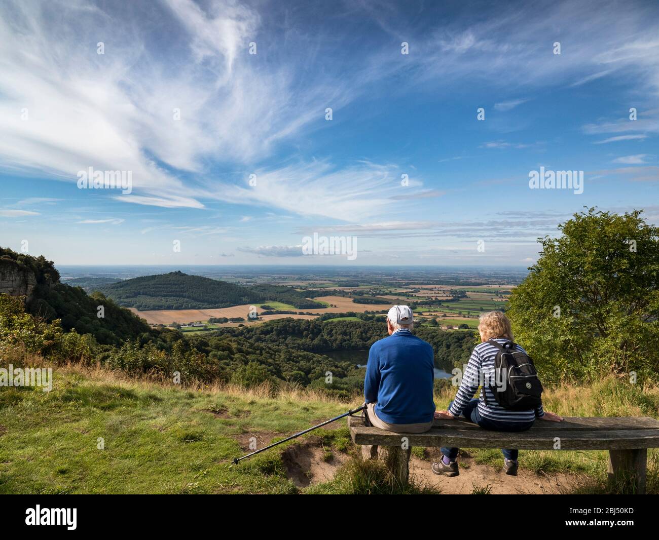 A old couple sat on a wooden bench looking over the Vale of York from Sutton Bank. Stock Photo