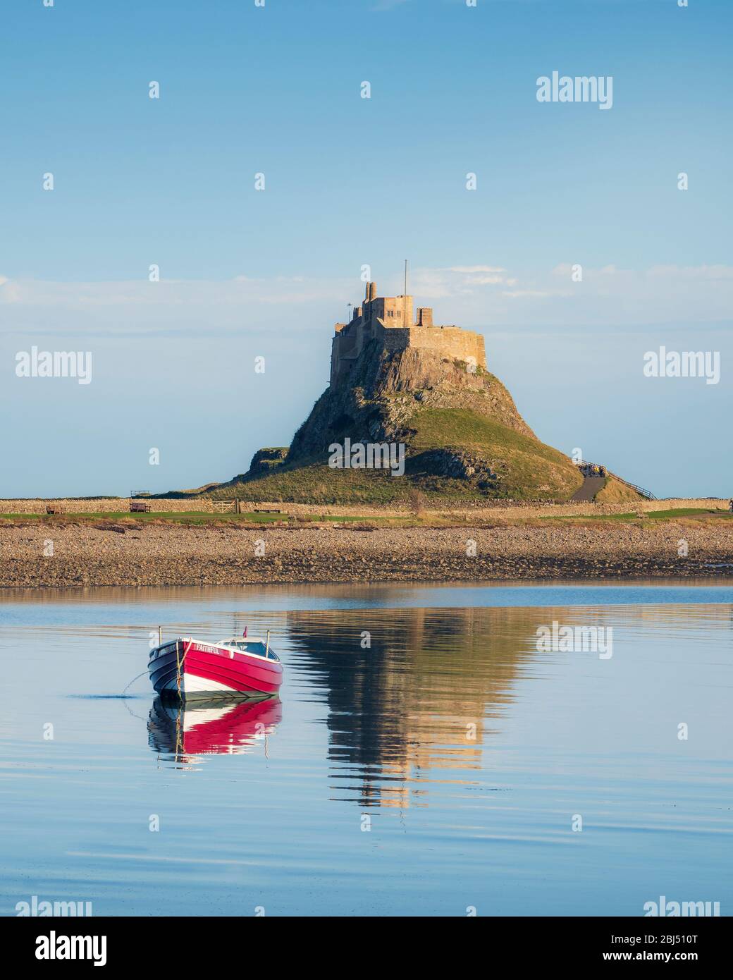 A small red boat in front of Lindisfarne castle reflecting in the sea. Stock Photo