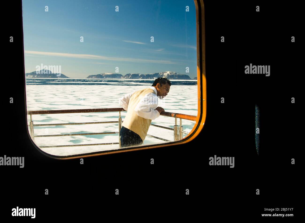 Smartly dressed Asian waiter seen through a porthole looking at icebergs on an expedition ship in the Arctic. Stock Photo