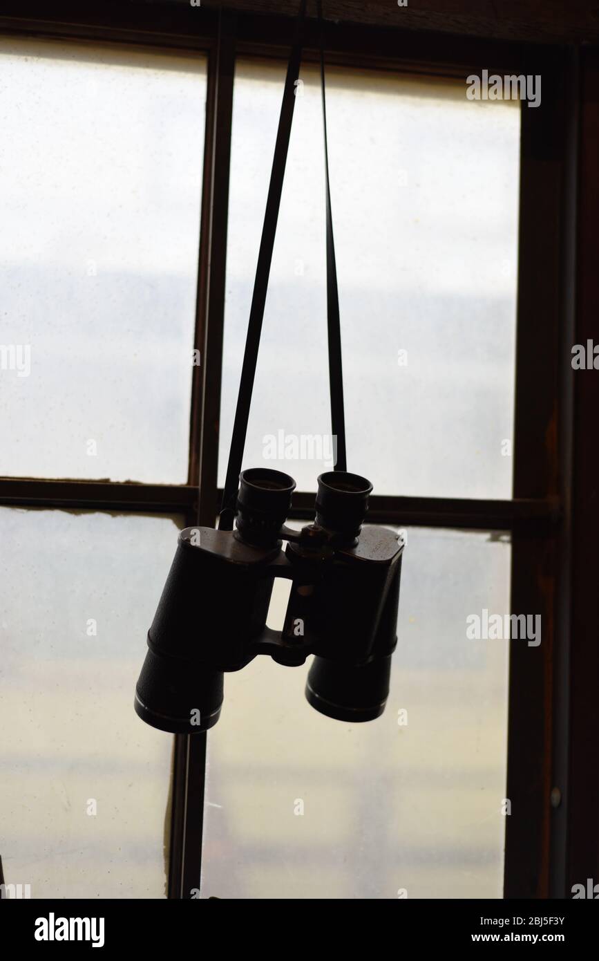 An old pair of binoculars hang from a strap in front of a dirty window Stock Photo