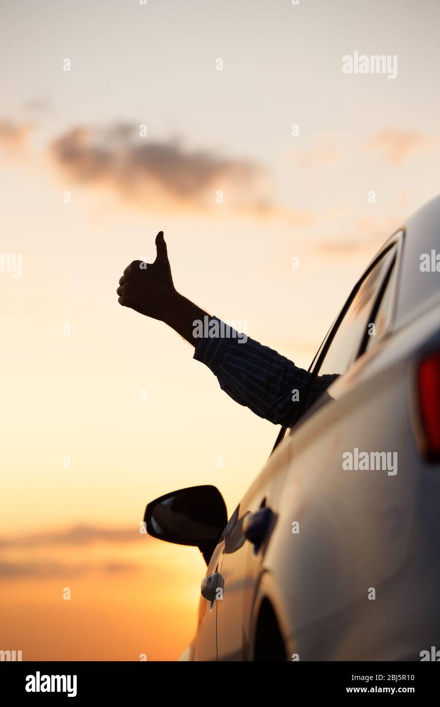 Man showing thumbs up/making Like / Ok sign with hand from car window with sunset sky, relaxing, enjoying road trip and feeling the air and freedom. T Stock Photo