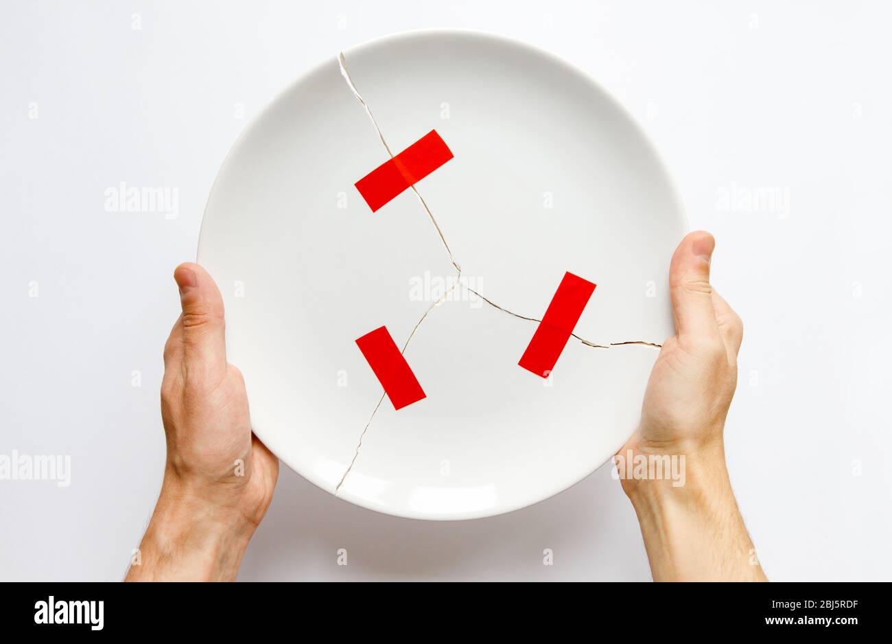 Top view of man hands holding a broken white plate, parts glued with red tape. Metaphor for divorce, relationships, friendships, crack in marriage. Lo Stock Photo