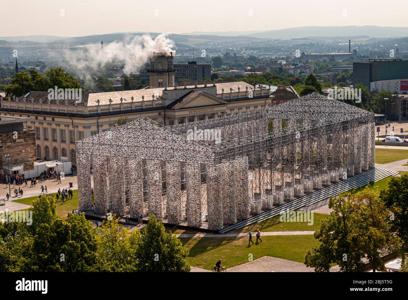 The Parthenon of Books, by Argentine artist Marta Minujin at documenta 14 in Kassel. Every day the construction, which in its dimension is a replica of the temple on the Acropolis, became more massive and filled with more books. In the background, the installation "Expiration Movement" smokes in the Zwehrenturm, Kassel, Germany Stock Photo