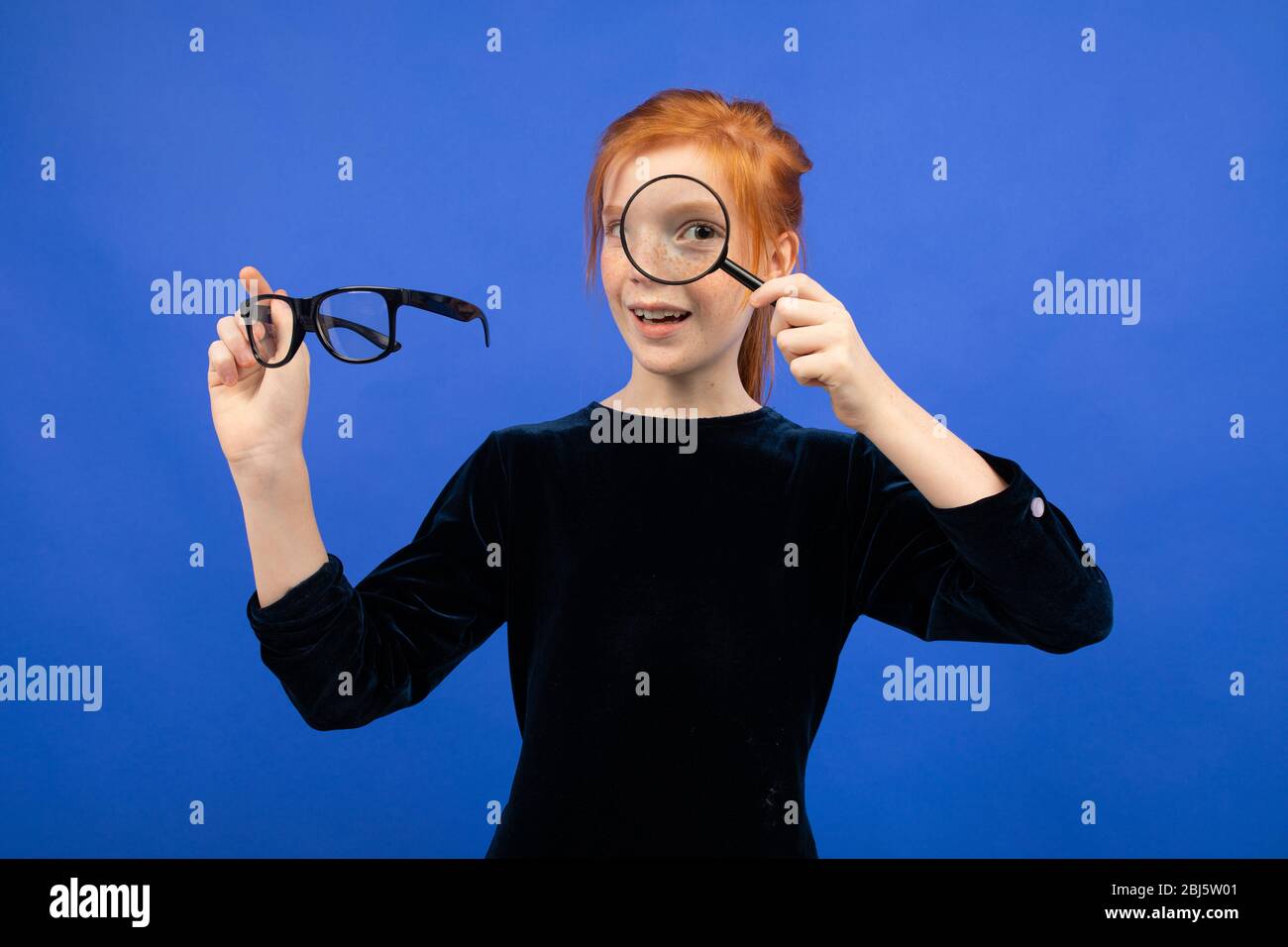 red-haired girl holding glasses for sight and a magnifier on a blue background. Stock Photo