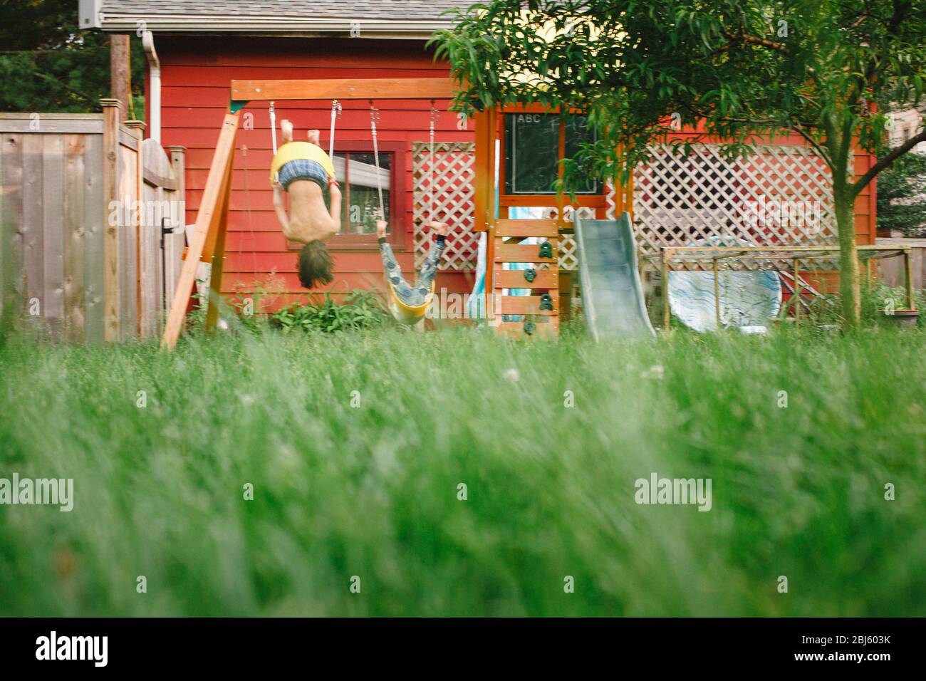 Two children flip upsidedown on a play set in their back yard Stock