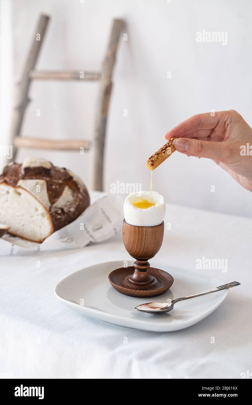 A hand soaks a cracker into the yolk of a broken boiled egg on a wooden stand on a table with a white tablecloth and bread in the background Stock Photo