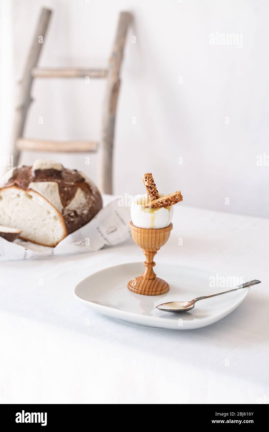 Two crackers with sesame seeds in a broken boiled egg on a wooden stand on a table with a white tablecloth and bread in the background Stock Photo