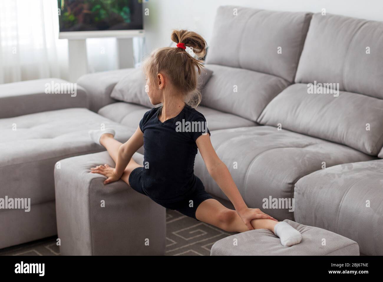 Little cute girl is practicing gymnastics at home. Online training. Stretching, twine. Quarantine. Stay at home Stock Photo