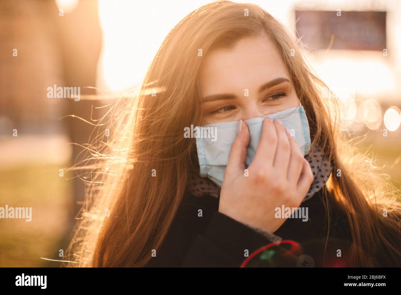 Portrait of happy young woman wearing protective face medical mask standing in city at sunset Stock Photo