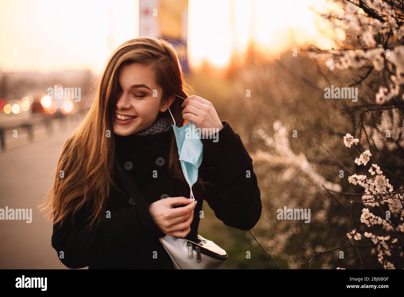 Happy cheerful young woman removing face medical mask while standing on street in city during sunset in spring Stock Photo