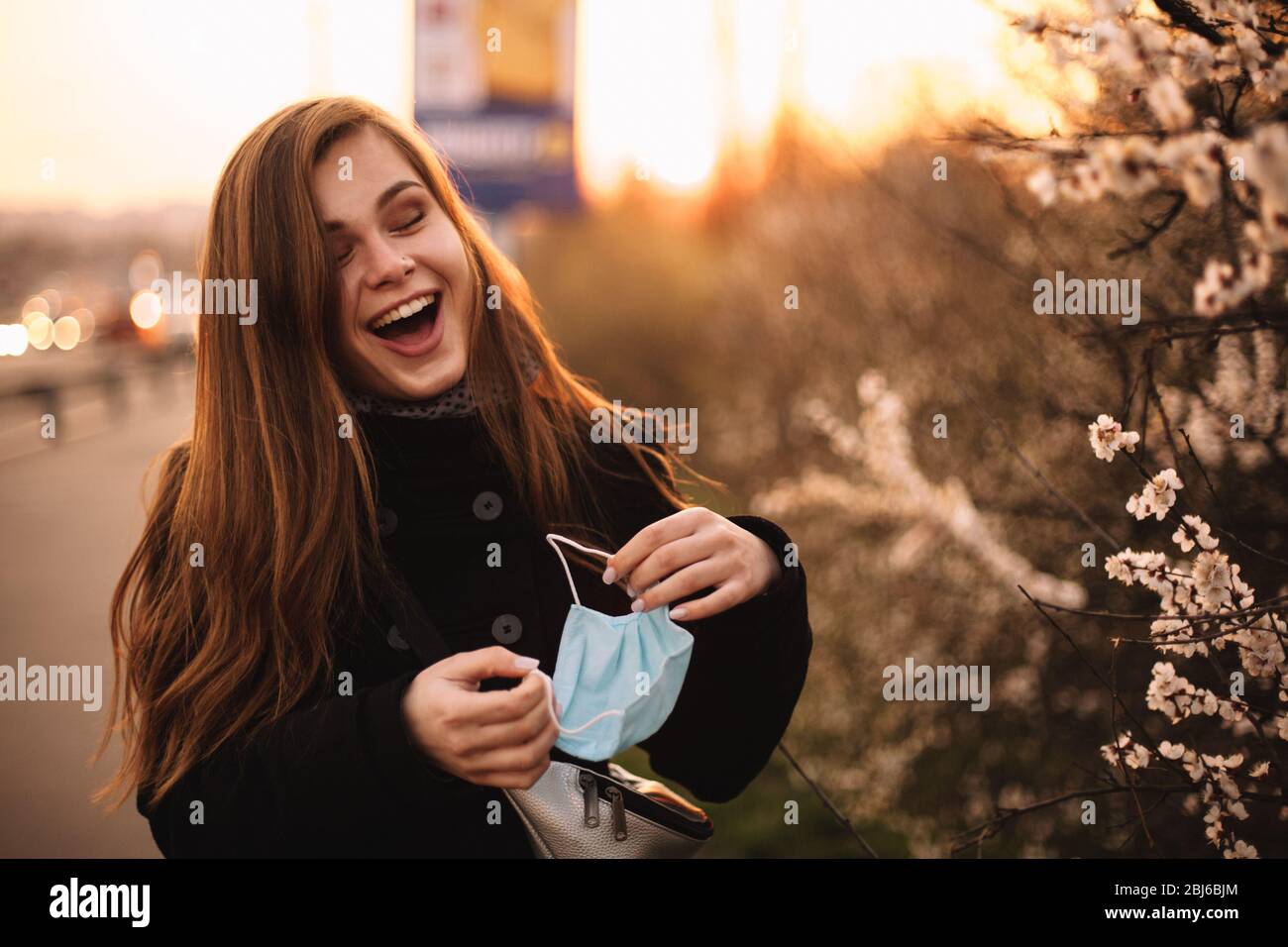 Happy cheerful young woman removing face medical mask while standing on street in city during sunset in spring Stock Photo
